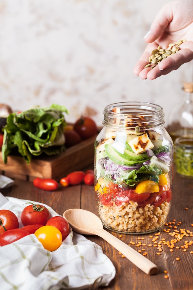 A healthy salad in a jar sits on a wooden table, with a hand gently resting beside it, showcasing fresh ingredients.